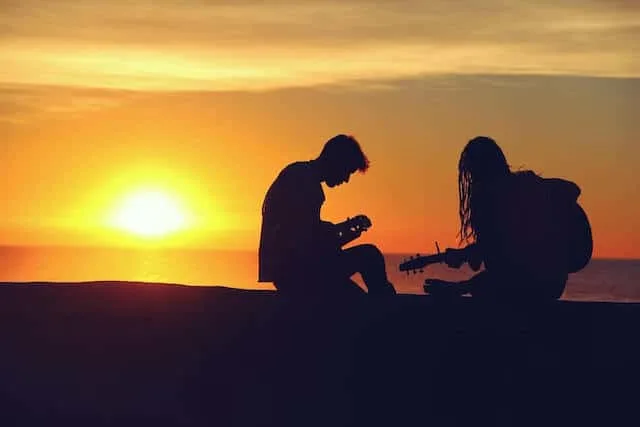 Silhouette on a Couple on the beach with the sun setting behind them. The woman on the right is holding a guitar, the man is sat looking at something in his hands
