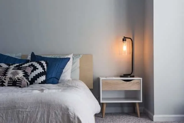 Half of a bed in a hotel room with white bedding and a navy blue accent pillow next to a wooden bedside table with a black filament bump lamp lit up on it