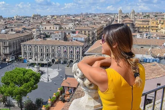 woman in a yellow sleevless dress with brown hair tied in a ponytail lookout our over the city of Catania in Sicily