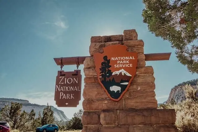 US National Park Service sign on a red brick wall with a hanging wooden sign saying Zion National Park