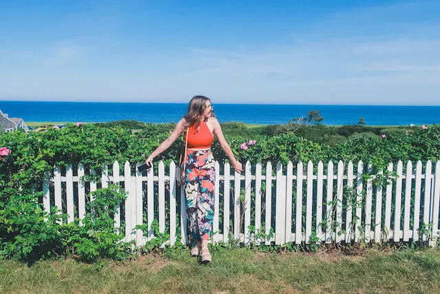 A woman leaning against a white picket fence with the ocean in the backgorund on Sconset Bluff Walk 