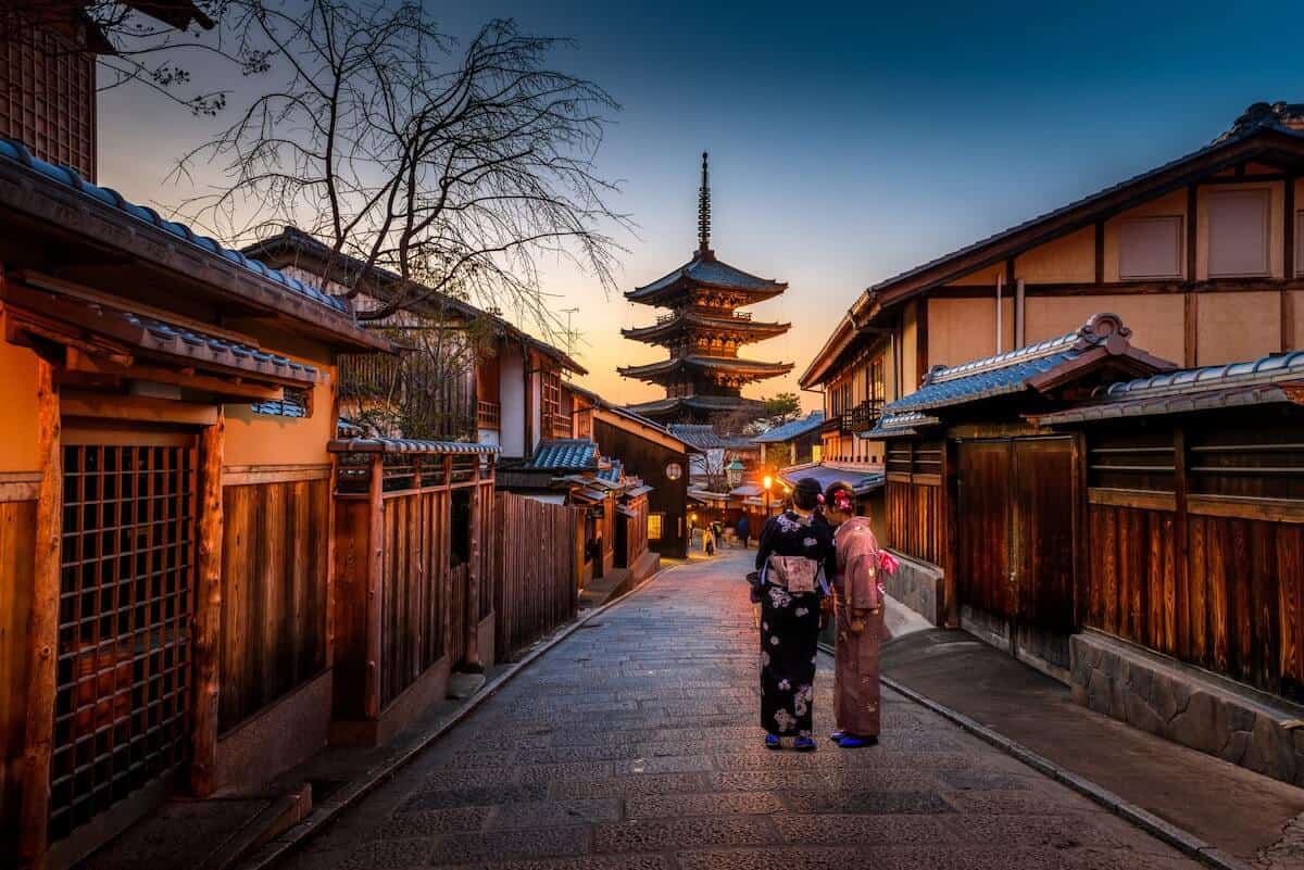 Header image for the Best Things to do in Japan of a narrow street lined with single storey houses, a 5 level pagoda in the distance and two women dressed in traditional kimono with a parasol in the foreground