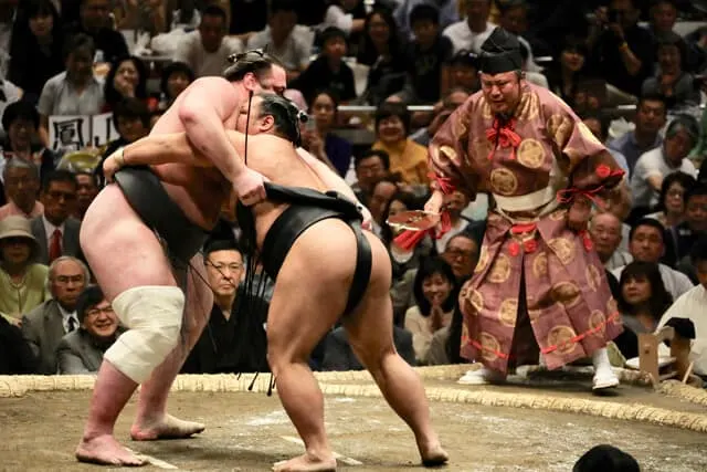 Two sumo wrestlers in the ring, a robed referee stands in the background and fans surround the ring
