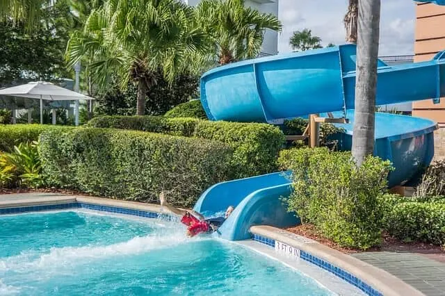 A blue water side tube twisting around into a pool flanked by green hedges either side with a child exiiting the slide feet first into the water