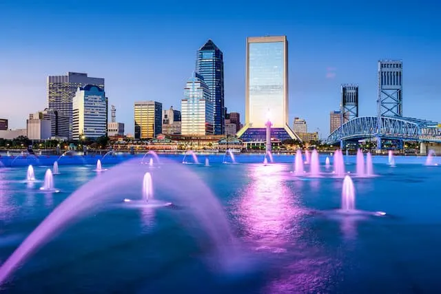 a lake with multiple lit up fountains glowing purple in front of the city sykline at dusk