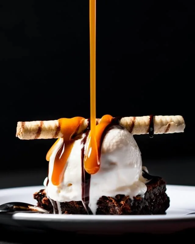 A chocolate brownie on a round white place with a scoop of vanilla ice cream on top, plus a twille and a drip of caramel sauce being poured over the whole thing