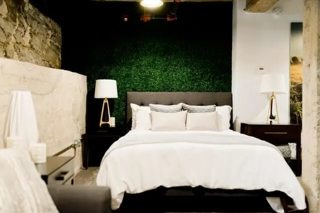 Hotel room with green grass textured wall behind a double bed with white bedding and off white throw cushions. A black bedside table sits on e right with a white lamp sitting on top of it
