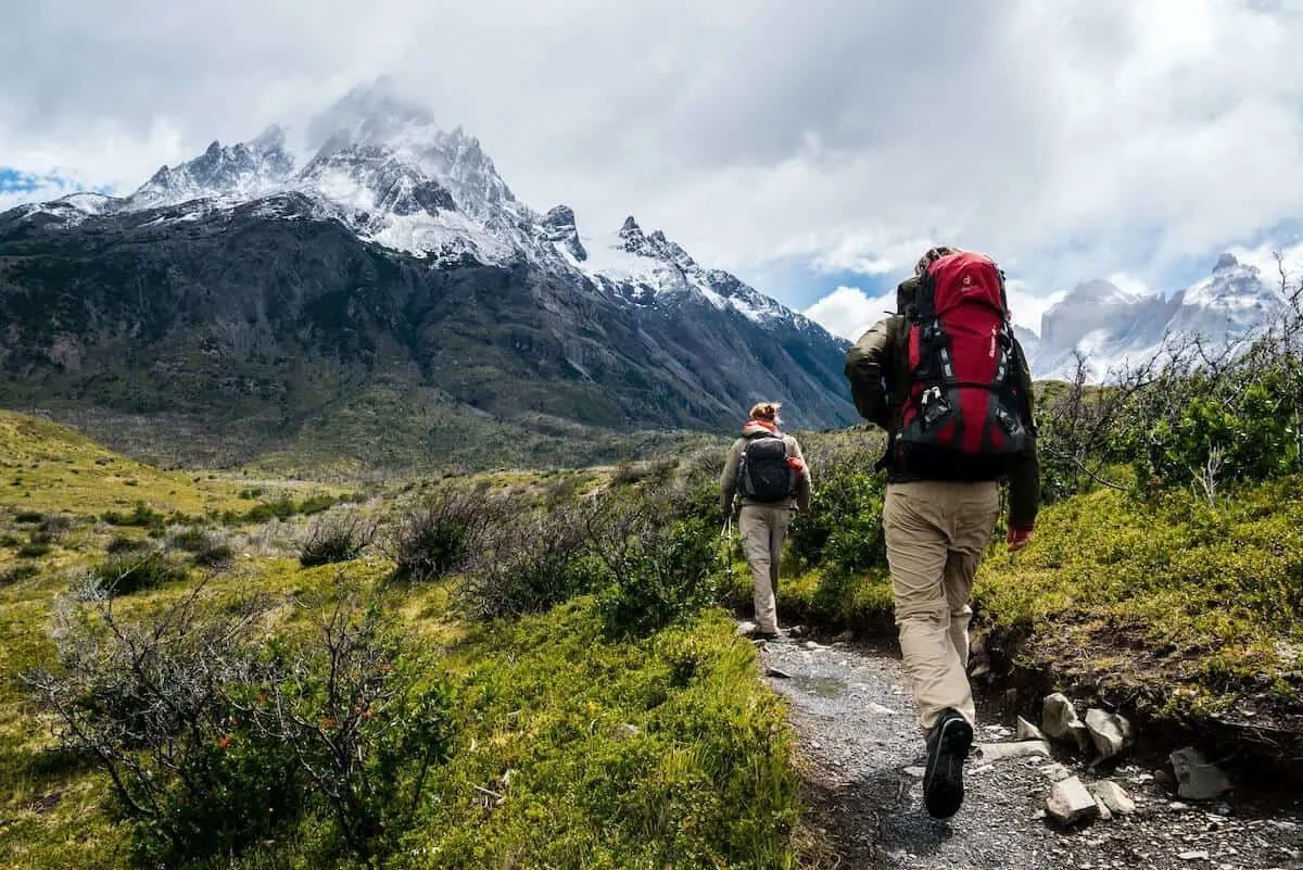 Hiking Tips for Beginners cover image of a couple hikers walking along a gravel path with grass either side walking towards a snow topped mountain in the distance