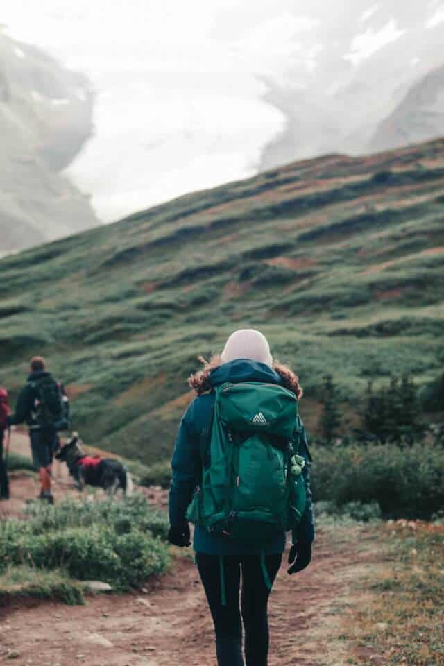 Woman wearing a green coast with fur hood carring a green backpack walking away from the camera along a hiking trail