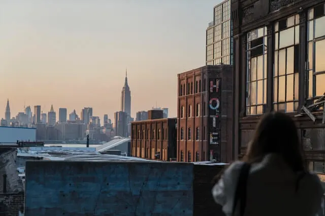 Woman standing on a balcony looking towards the new york city skyline, a building directly in front has the word 'Hotel' in capital letters down the side