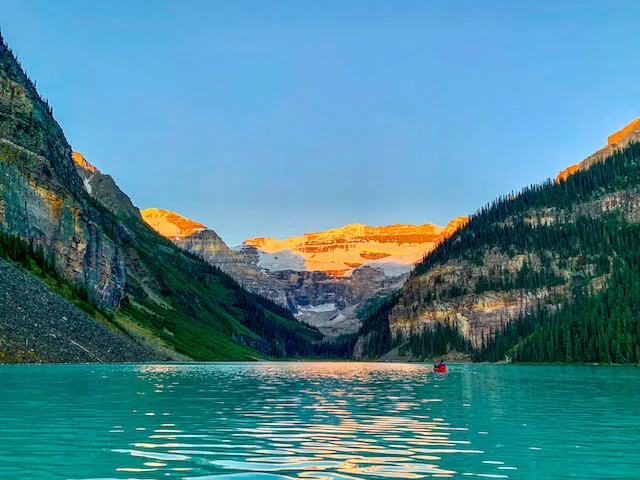 Blue water of Lake Louise at sunrise, a person in a red canoe in the distance towards the mountains of the background