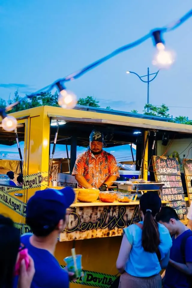 Food truck, male server behind the counter, festoon lights hanging around the site, 2 people waiting in front of the truck for food