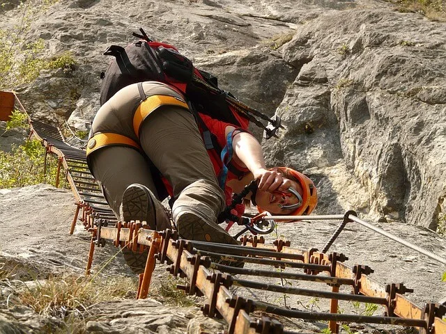 Woman climbing up a steel ladder built into the cliffside wearing hiking pants, a backpack, red tshirt and crash helmet