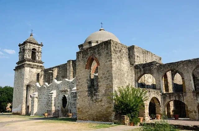 Old, ruined, discoloured white stone mission building with central domed roof and bell tower with arched window spaces