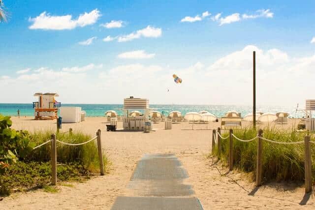 wooden walkwal part covered in sand leading to the beach with sun loungers spead out on the sand and the blue ocean in the background on a sunny day