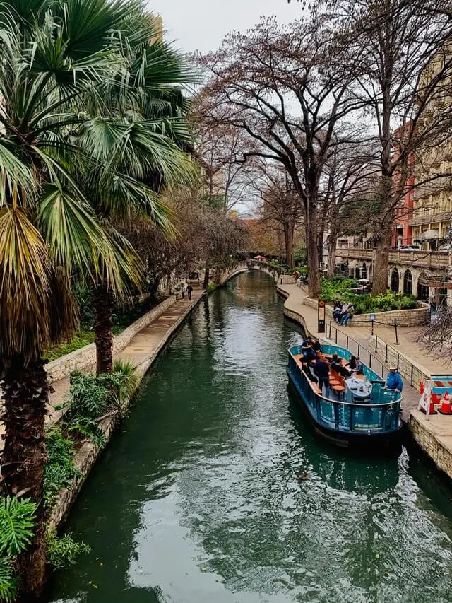 Central river with a stationarary boat docked o the right, paved pathways either side of the water which is flanked by palm trees.