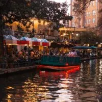Fun Things to do in San Antonio cover photo of the Riverwalk at dusk with people sitting at a bar under umberellas next to a parked boat.