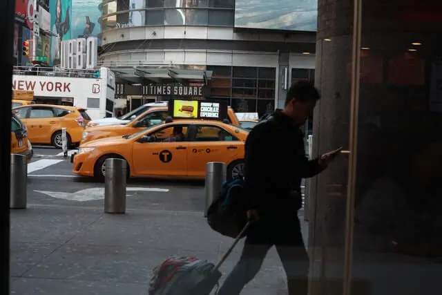 Yellow Cabs on the street in the background with a man pulling a wheeled suitcase into a building while looking at his phone