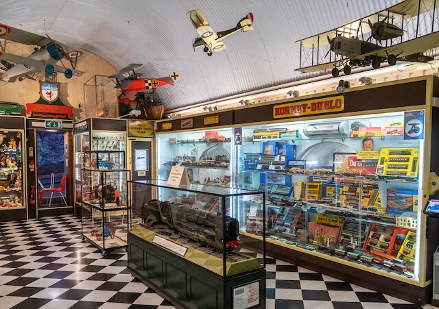 Inside Brighton Toy and Model Museum with checkerboard black and white floor, glass diplay cases of different toy brands and model airplanes hanging from the ceiling