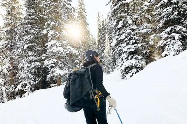 Camping in WInter - woman facing away from the camera hiking with ski poles dressed in snow gear with snow covered fir trees in the background
