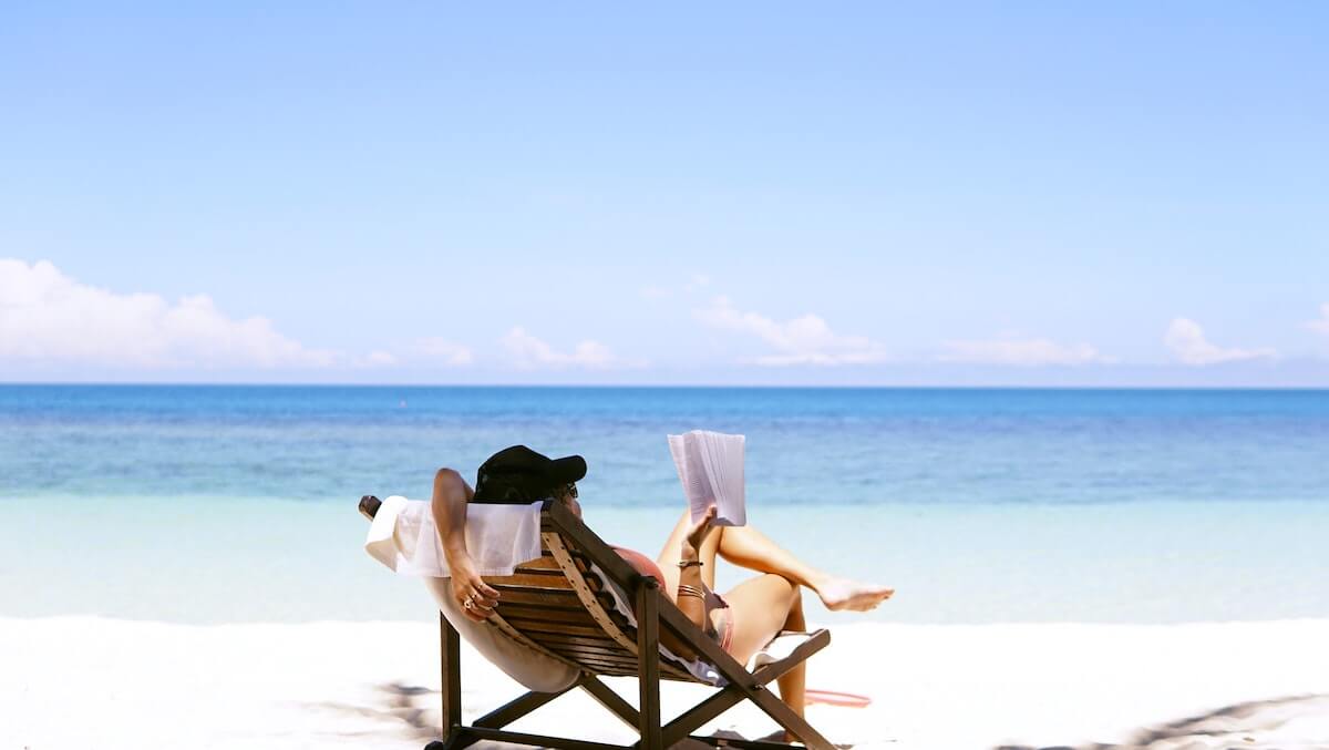 Vacation Quotes & Vacation Captions for Your Next Trip header photo of a woman sitting in a deck chair on the each facing the ocean behind her and reading a book wearing a sunhat and bathing suit