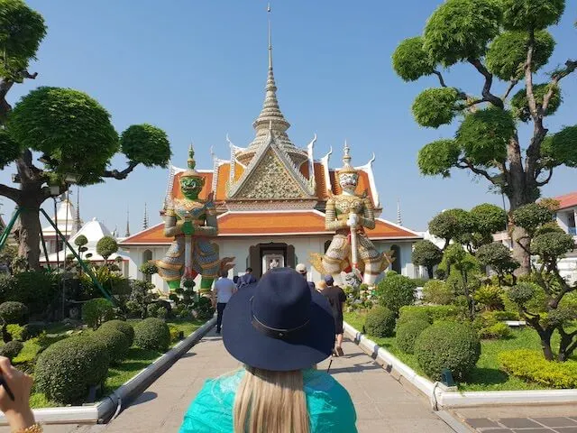 Woman in a blue hat and a green shawl standing in front of a gold temple in Bangkok