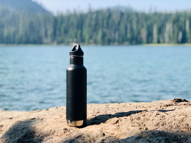 Black travel water bottle in focus on a rock in front of a lake, with a forest in the background.