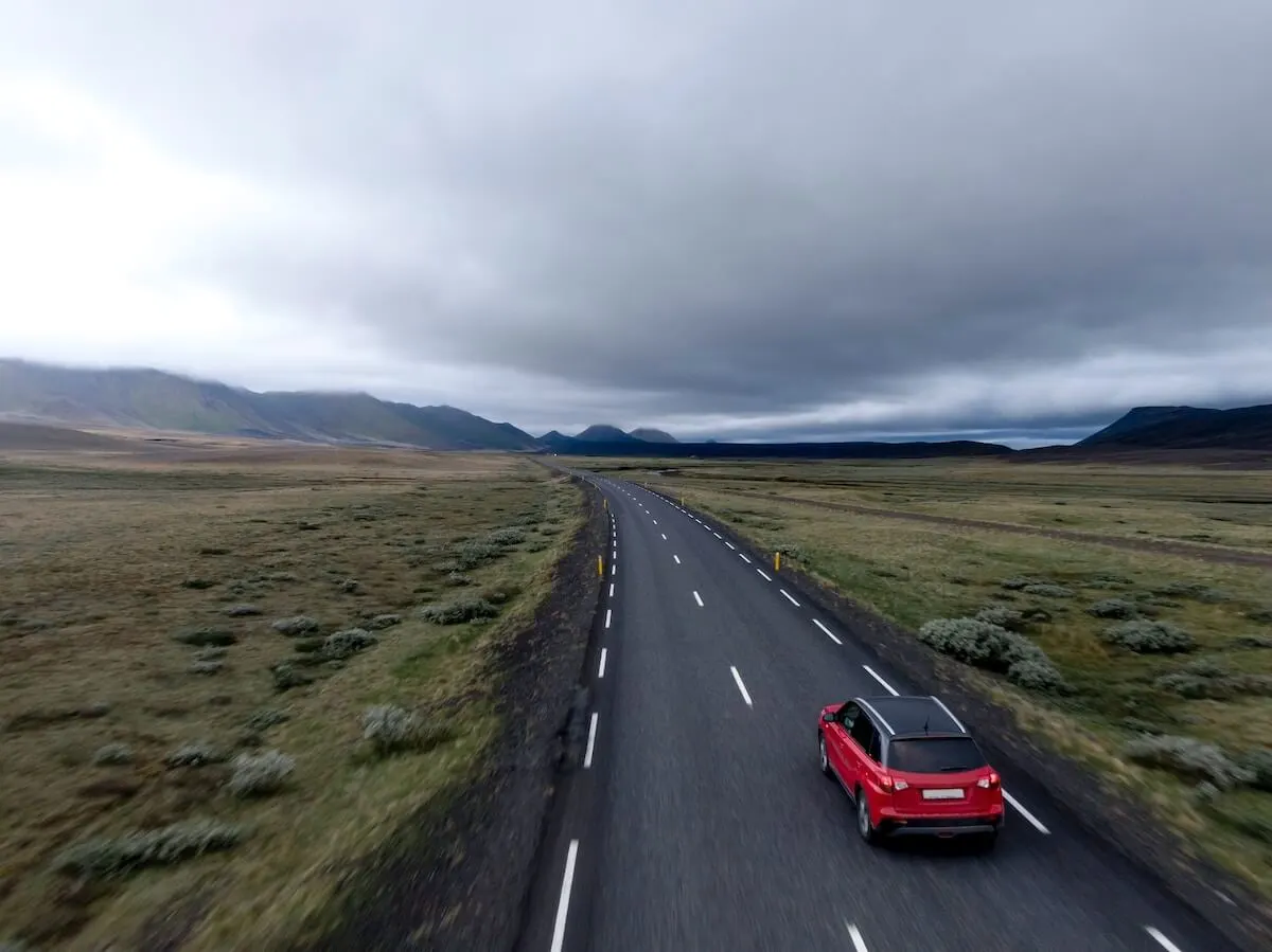 Money Saving Road Trip Tips cover photo of a red car driving on a black tarmac highway with grassy verges either side