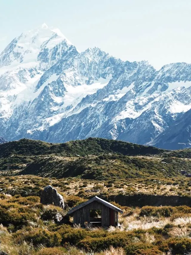 Small Wooden Mountain hut surrounded by scrubland in front of a huge snow covered mountain