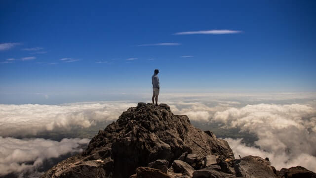 Man standing on top of a mound of dark rocks elevated above the cloud line at Mount Taranaki