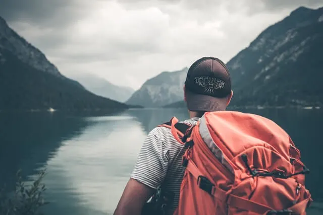 Man with a lrage orange backpack on his back, wearing a backwards facing baseball cap with his back to the camera looking a a lake surrounded by mountains