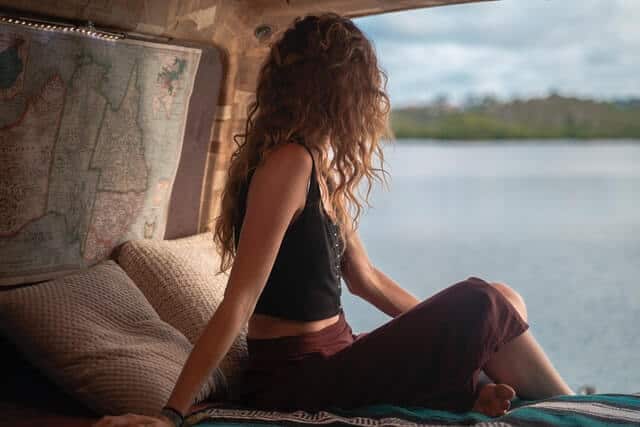 Long haired woman sitting in a campervan with the back doors open next to a lake
