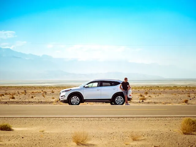 White SVU parked on the side of a sandy higway, a man in denim shorts and black tshirt leans on the back looking into the distance