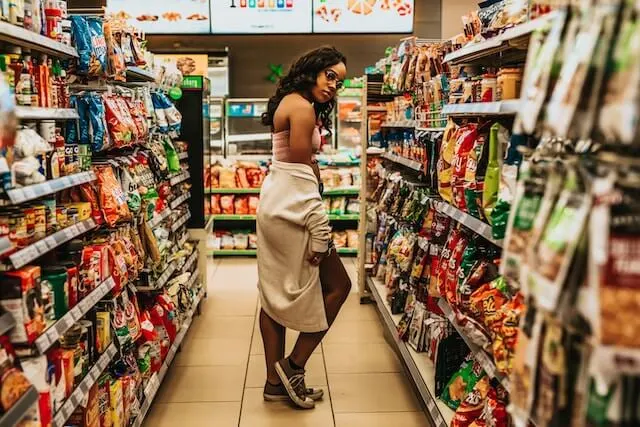 Woman standing between 2 aisles in a gas station, surrounded by snacks. She is wearning denim shorts, a while hooded top draped around her waist and a pair of blue converse