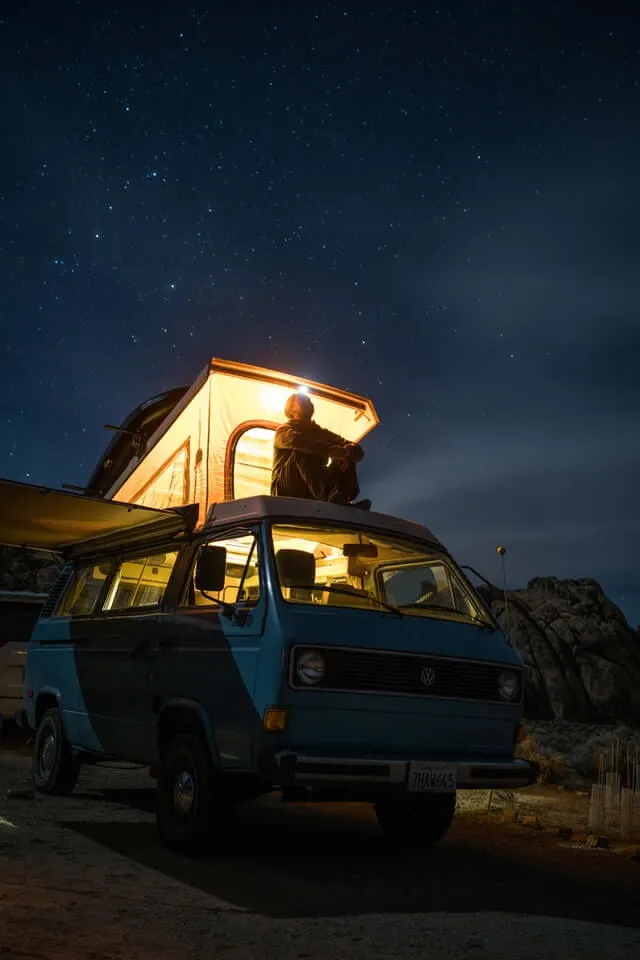 Man sitting on the roof of a campervan, with a pop up tent open behind him, wearing a headlamp, looking to the sky under a stary sky