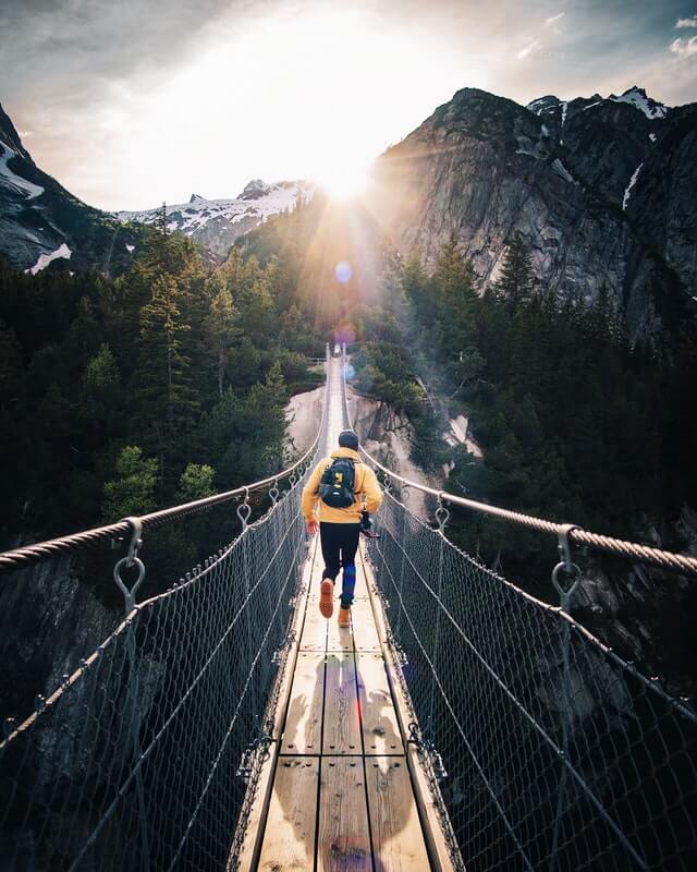 Man wearing a yellow coat, blue jeans and black hat, running across a wooden suspension bridge with wire sides