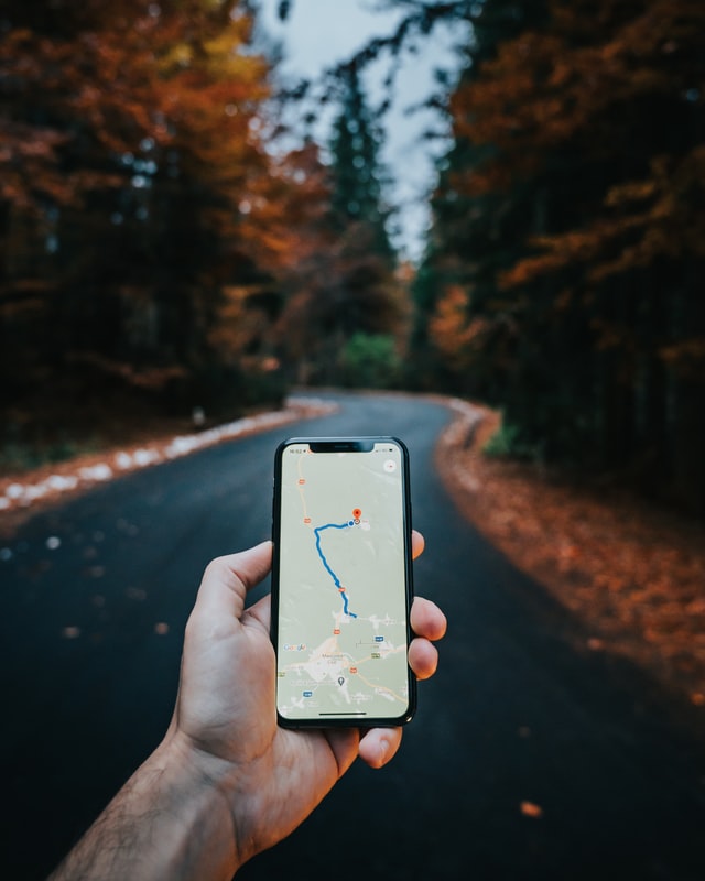 Mans hand holding a phone with a map and directions on the screen in focus in front of a road winding through a forrest