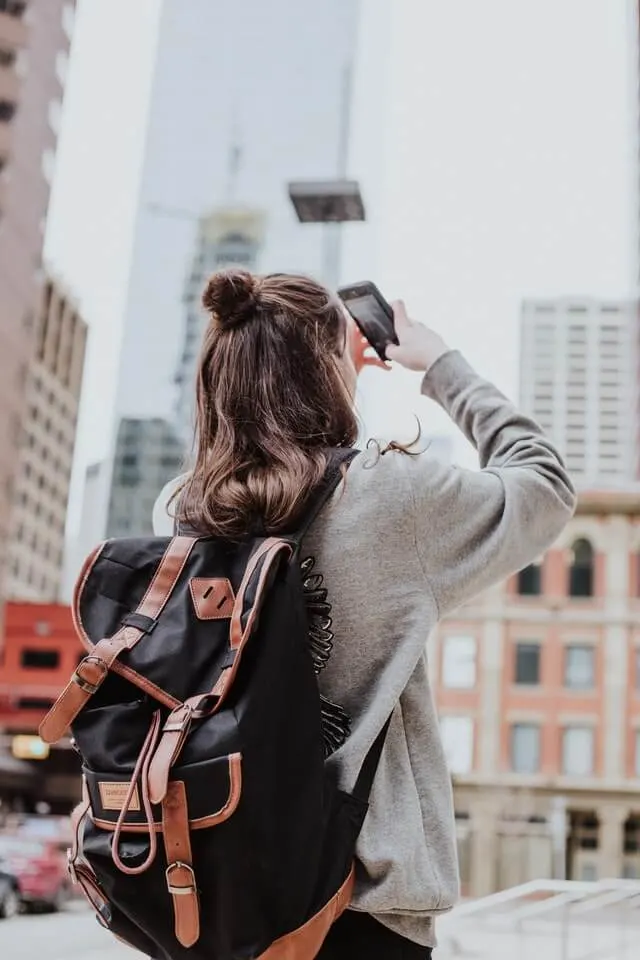 Woman wearing a grey jumper and black backpack, taking a photo with her iPhone