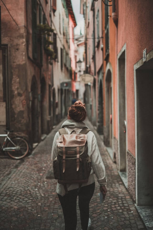 Woman with her hair tied in a bun wearing a backpack, white long sleeve shirt, black jeans, walking away from the camera on a narrow cobbled street with buidlings either side