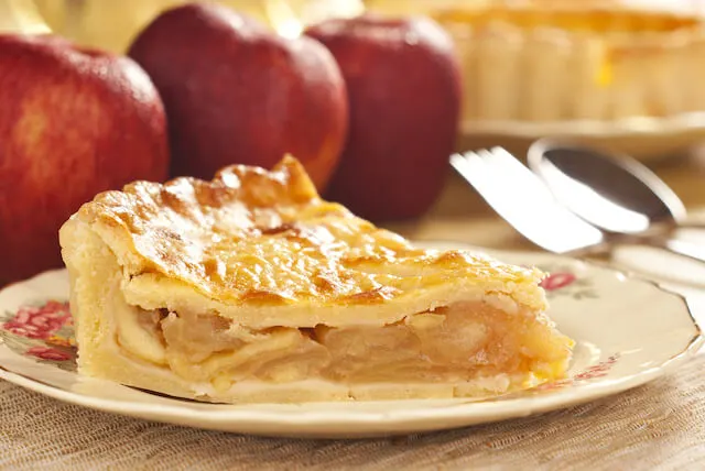 Slice of Apple pie on a plate in front of three red apples