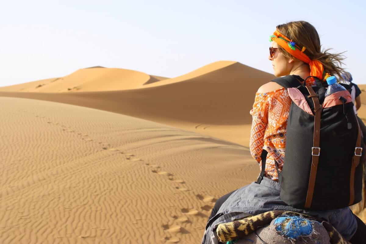 Solo Female Travel Tips cover photo of a woman wearing a backpack sitting on the back of a camel in the sand dunes