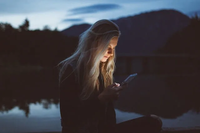 Woman looking at her phone, the brightness illuminating her face while sitting by a lake
