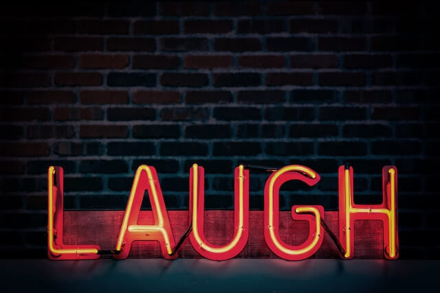 Neon sign in red saying 'Laugh' in front of a black brick wall