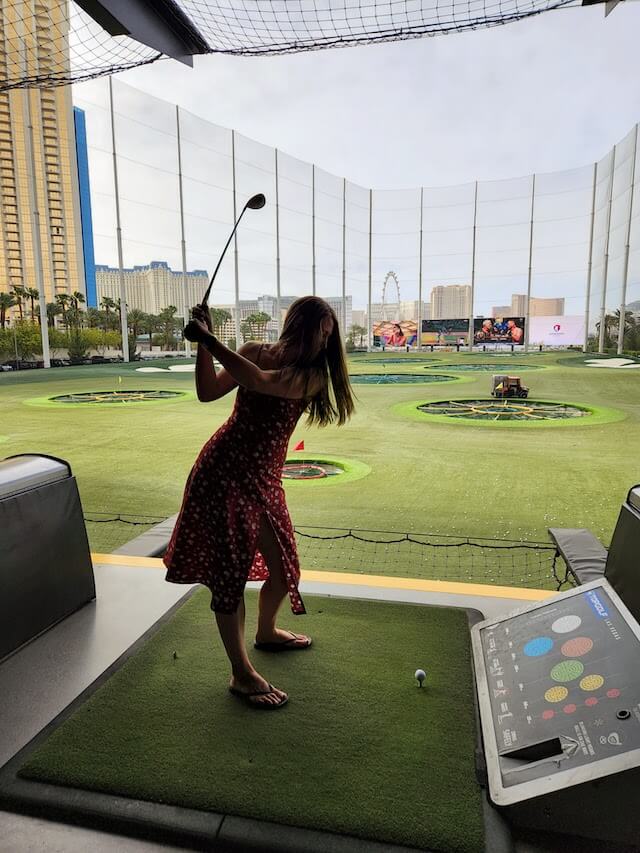 Woman in a red dress with long brown hair, swing a golf club at a driving range facility, the tall nets in the distances around the green playing area