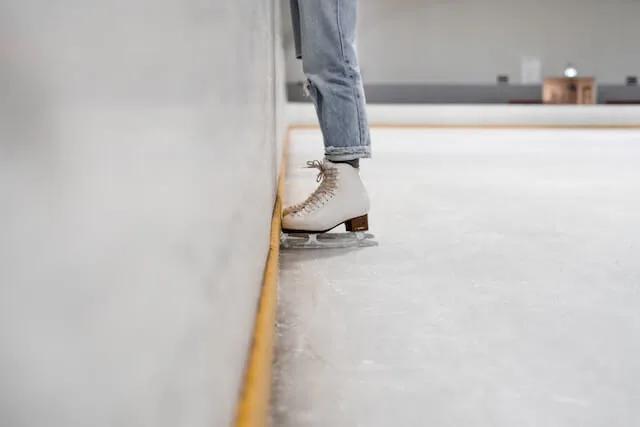 Woman wearing jeans and white skates standing facing the wall on an ice rink