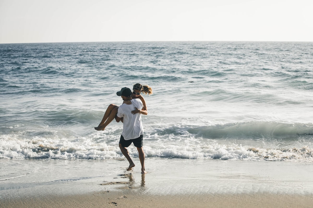 Romantic Getaways in California cover photo of a man carrying a woman on the beach near to the waters edge