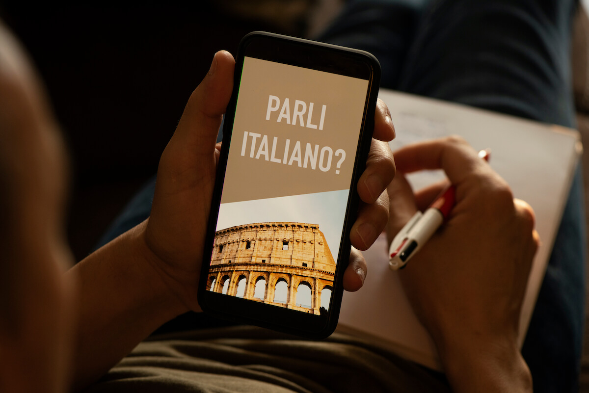 Basic Italian Words for Travel cover photo of a person holding a smartphone with a picture of the Colosseum and the words Parli Italiano on the screen