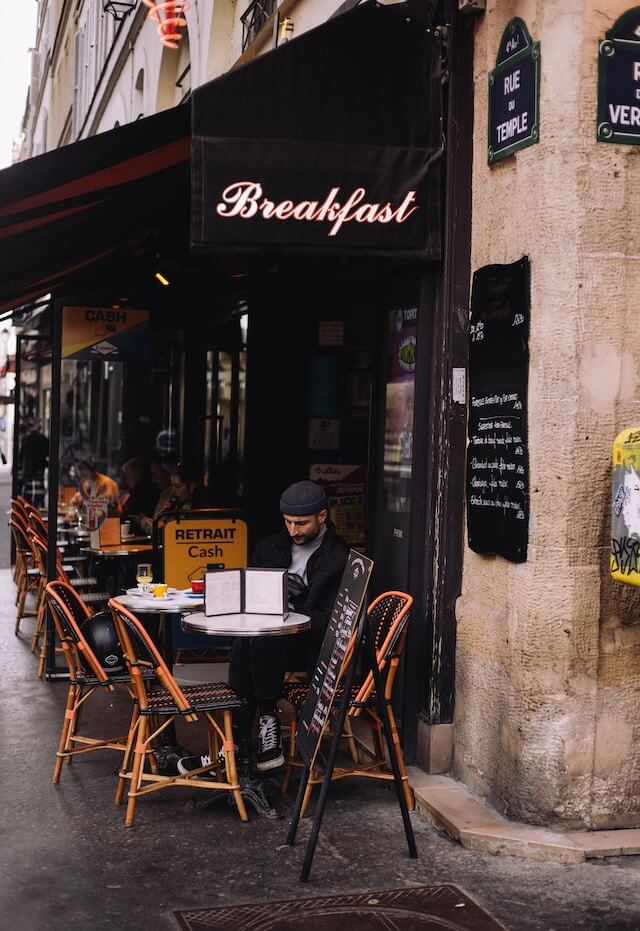 Man sitting outside a cafe under a sign that says breakfast