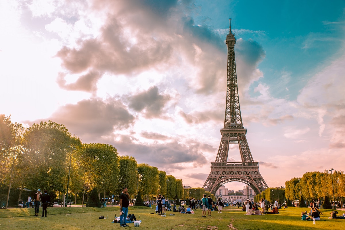 Movies Set In Paris cover photo of the Eiffel Tower and the park in front surrounded by a sunset pink and blue sky