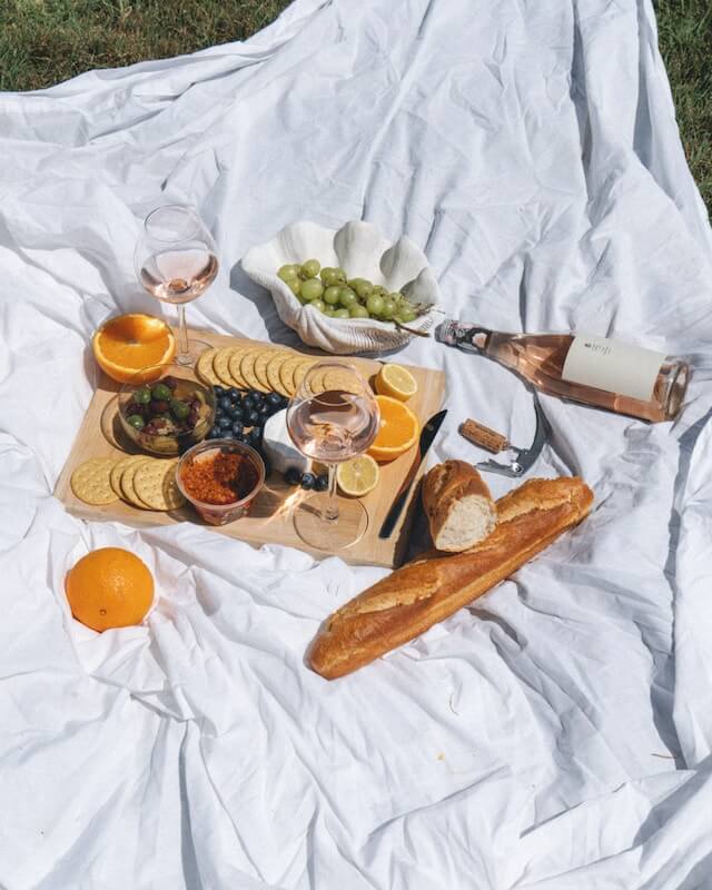 Pinic laid out on a white sheet including cheese, dips, grapes, baguette and rose wine